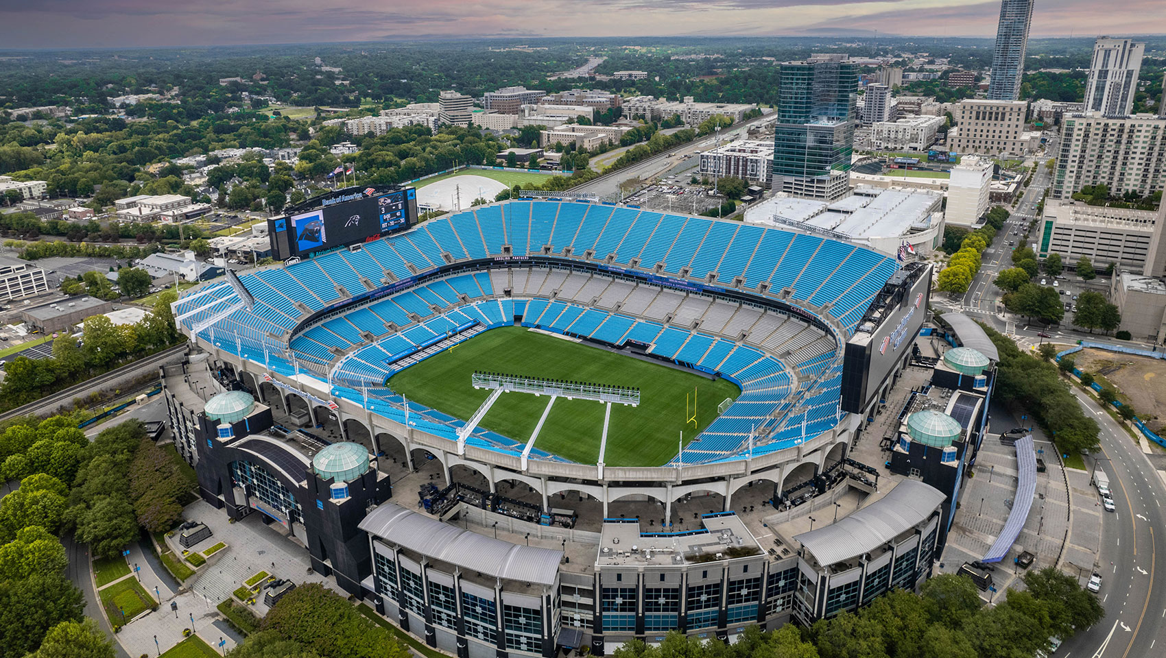 Aerial view of Bank of America Stadium in Charlotte, featuring blue seats, a green field, and the city under a colorful sky